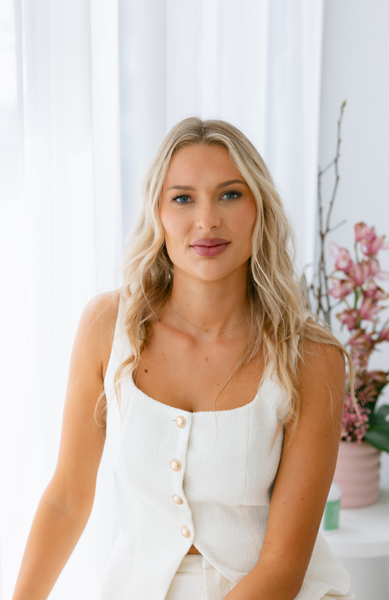 Woman in a white sleeveless top sitting in a bright room with white curtains and pink flowers.
